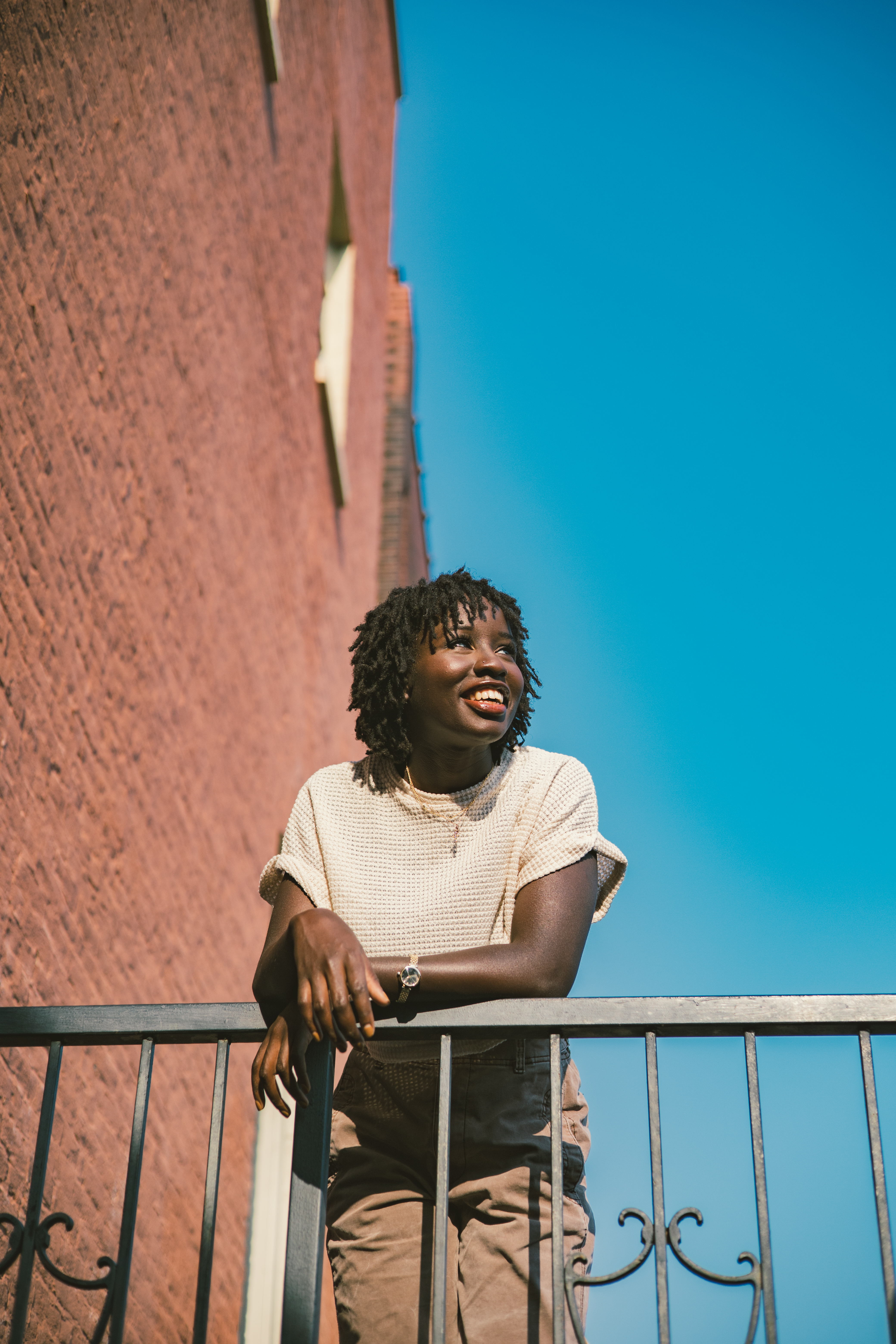 Young woman with dreadlocks smiles on a balcony against a brick building and blue sky