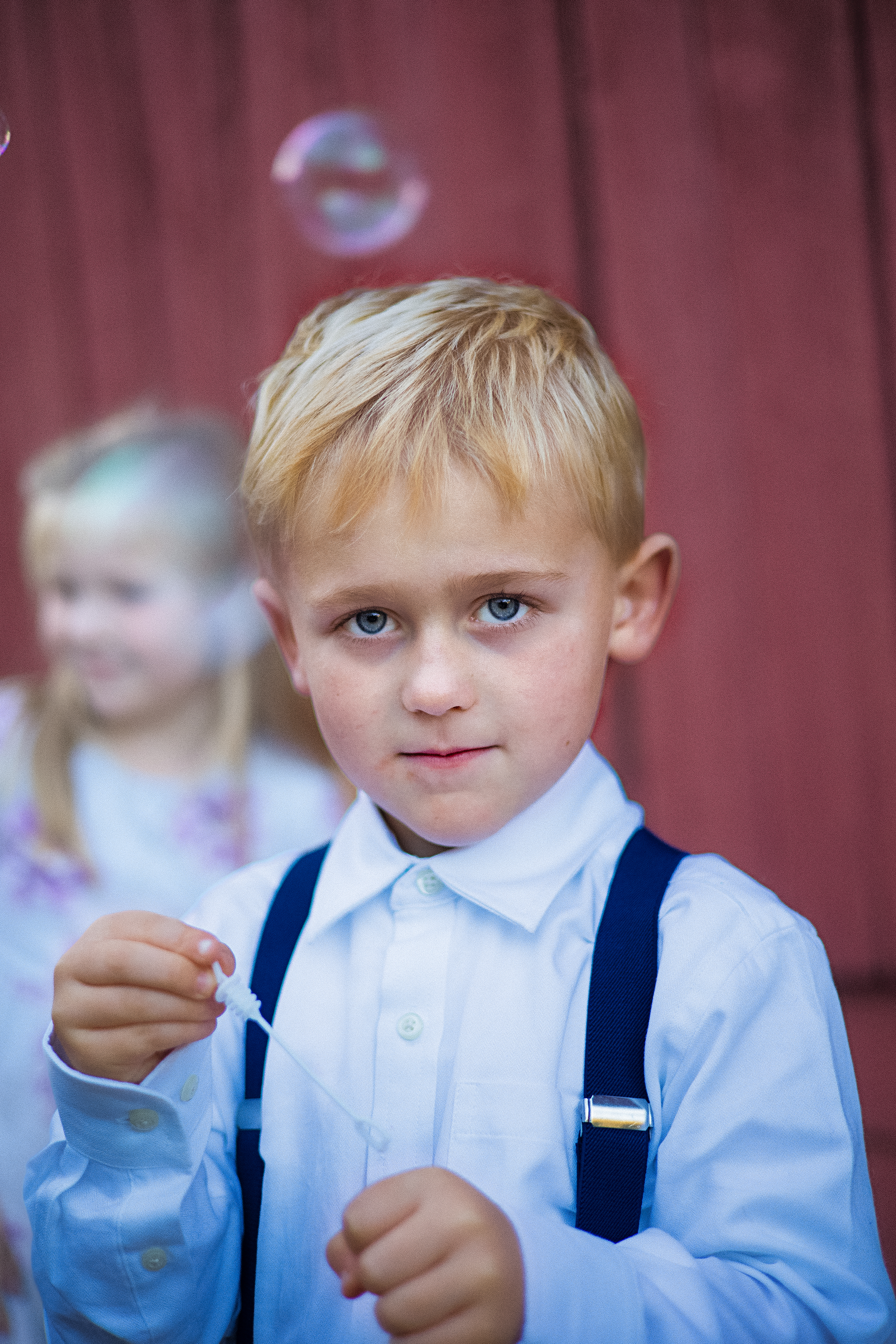 Young boy in suspenders blows bubbles at a wedding with a girl blurred in the background