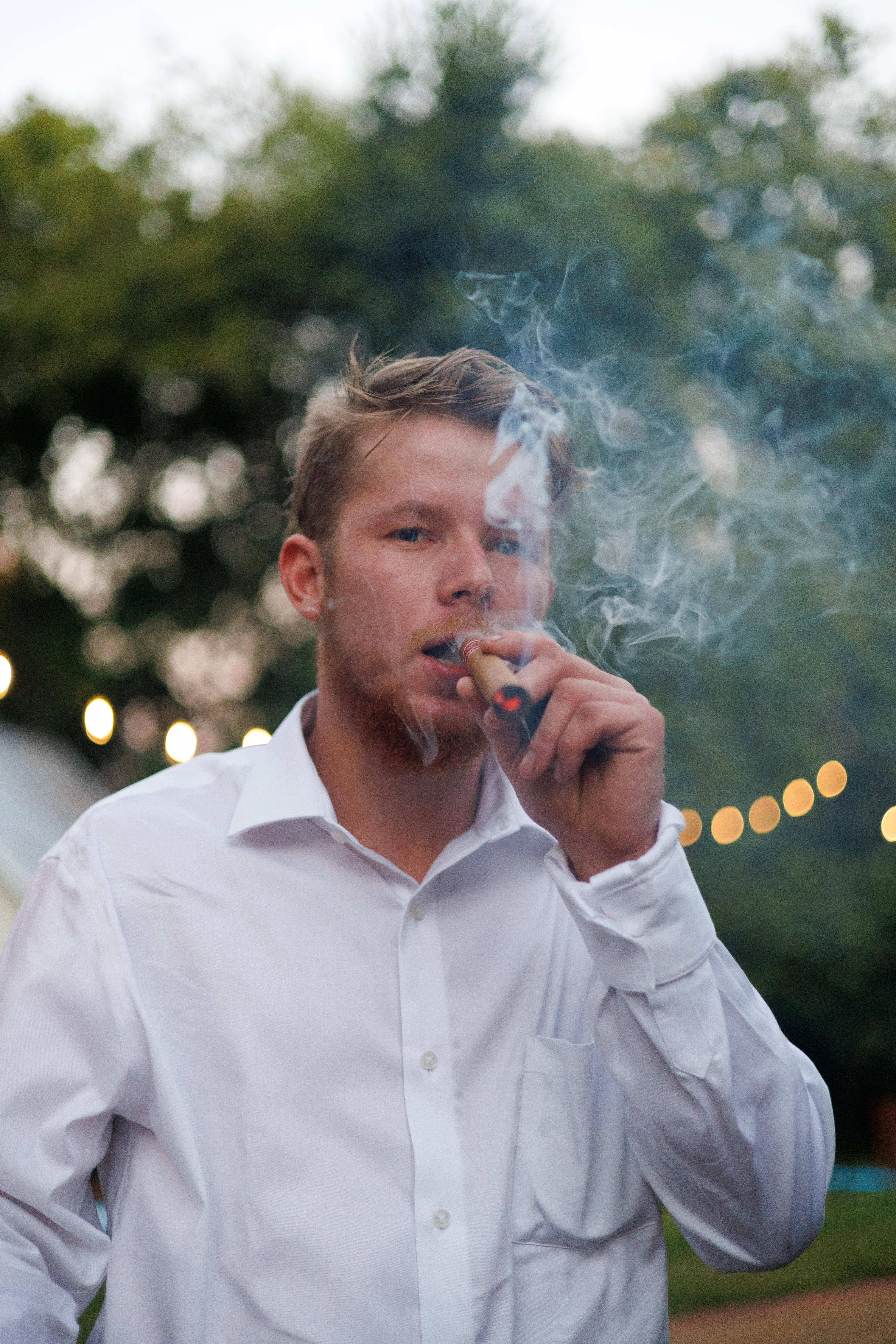 Groom smoking a cigar outdoors at a wedding with bokeh lights in the background