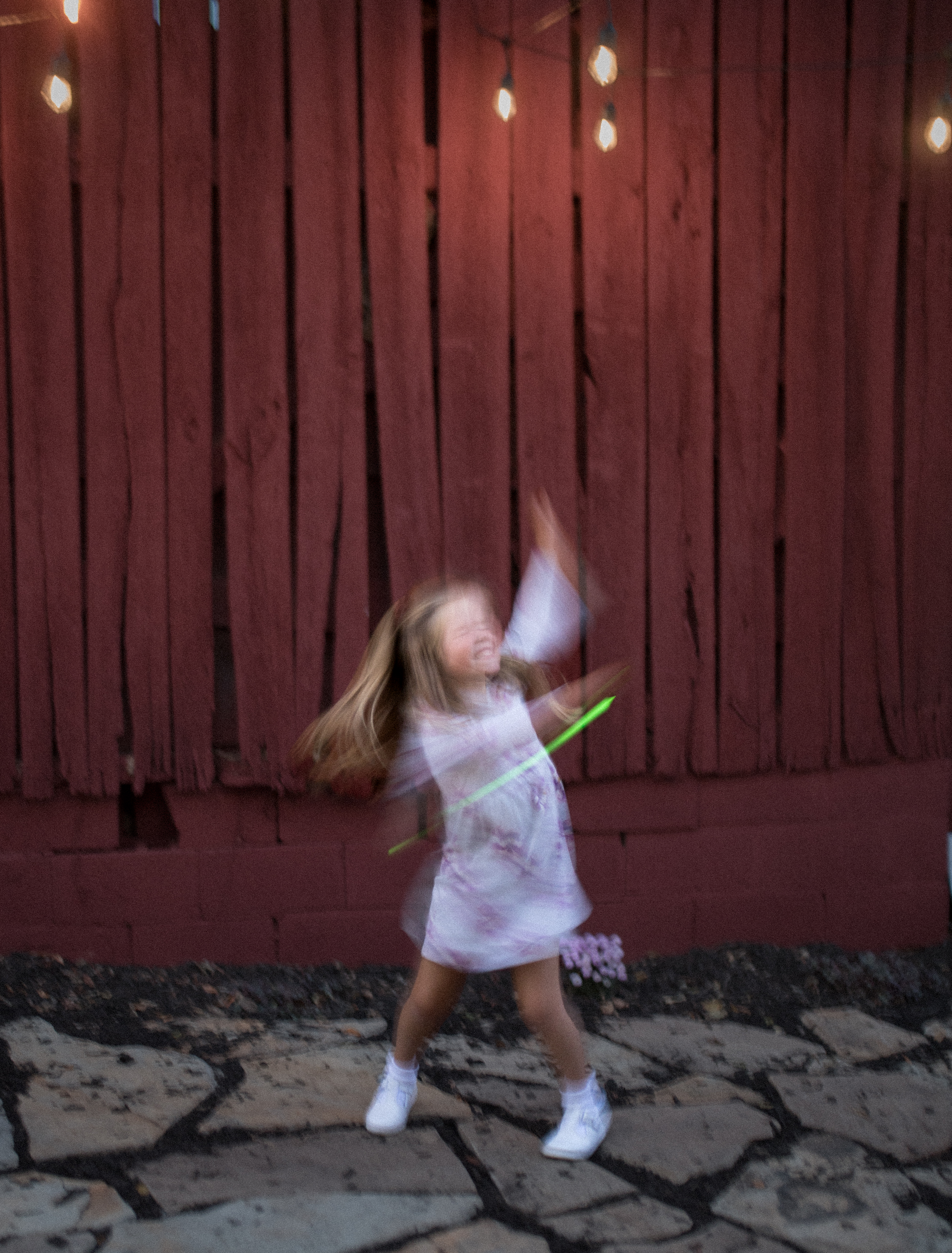 Joyful child twirls with glow sticks in front of a rustic wooden wall and string lights