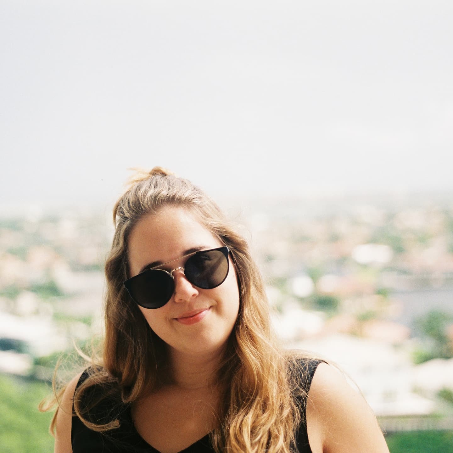 Young woman with sunglasses smiles outdoors with a blurred cityscape behind her