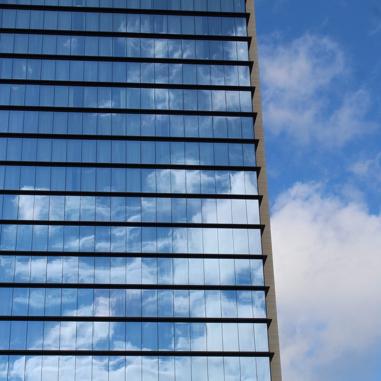 Reflective glass building facade mirrors a bright blue sky with scattered white clouds