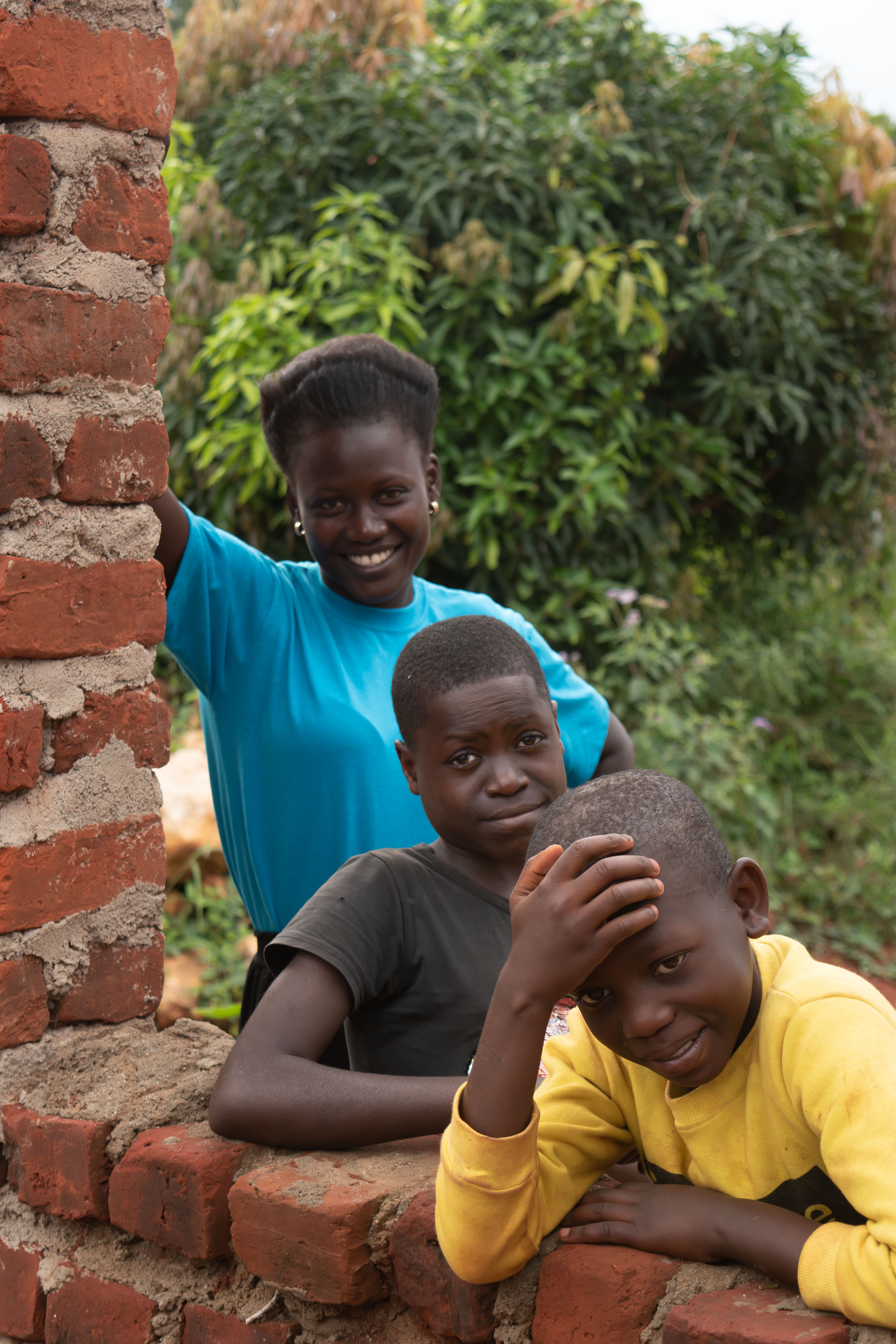 Three children smile and pose outdoors near a brick wall and lush greenery