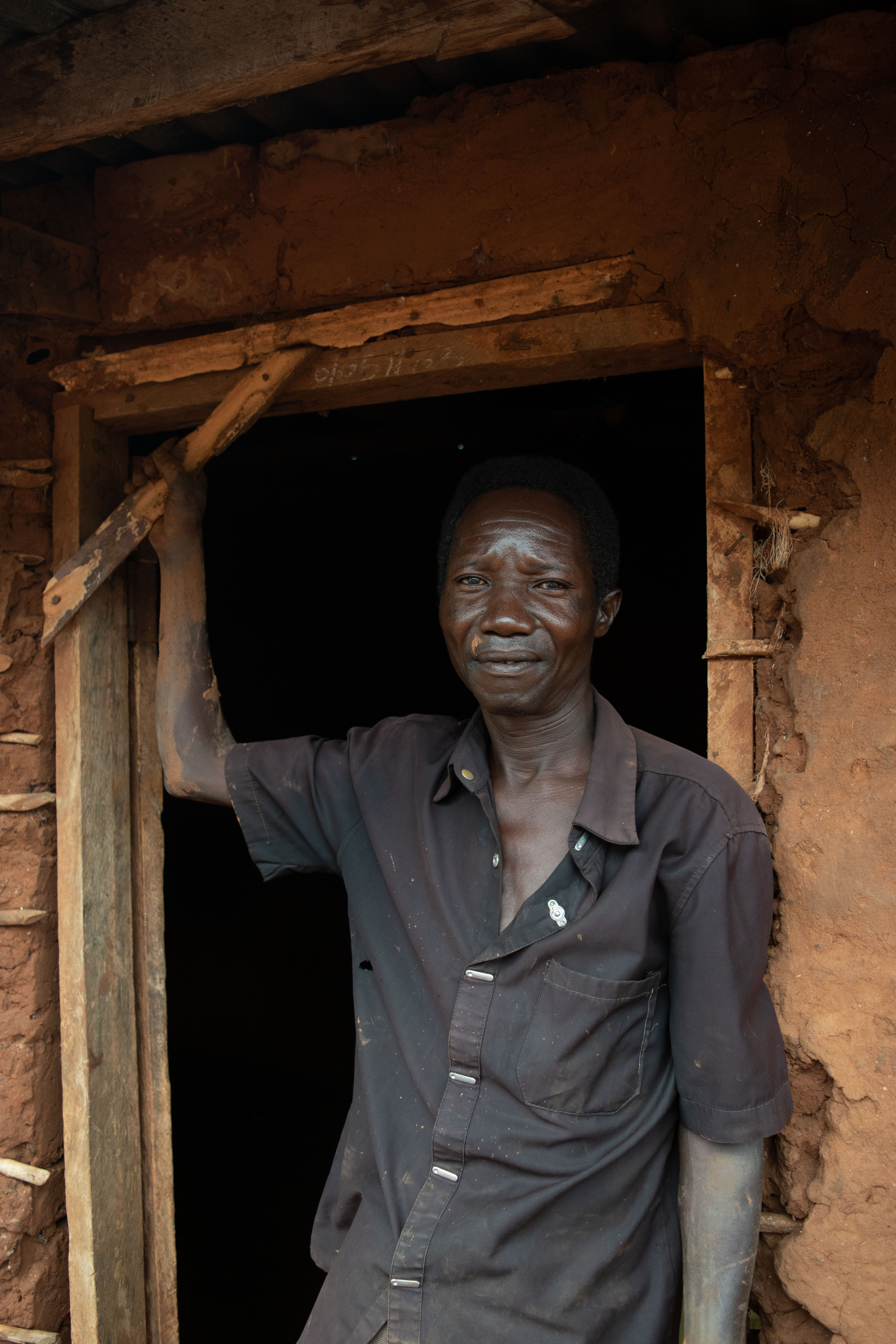 Man smiles, leaning on doorway of rustic mud home, natural light
