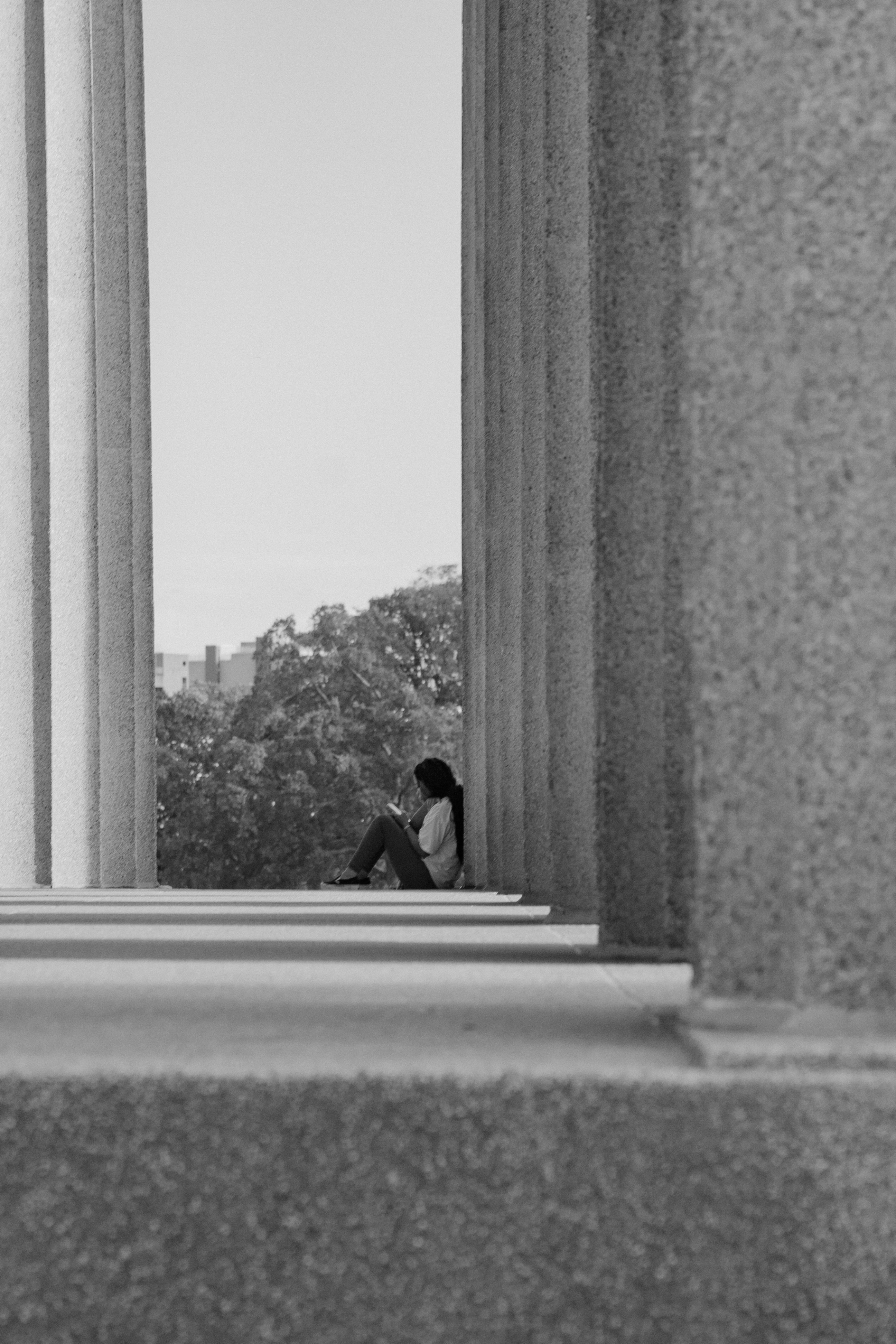 Woman sits between columns reading on her phone with trees and buildings in the background