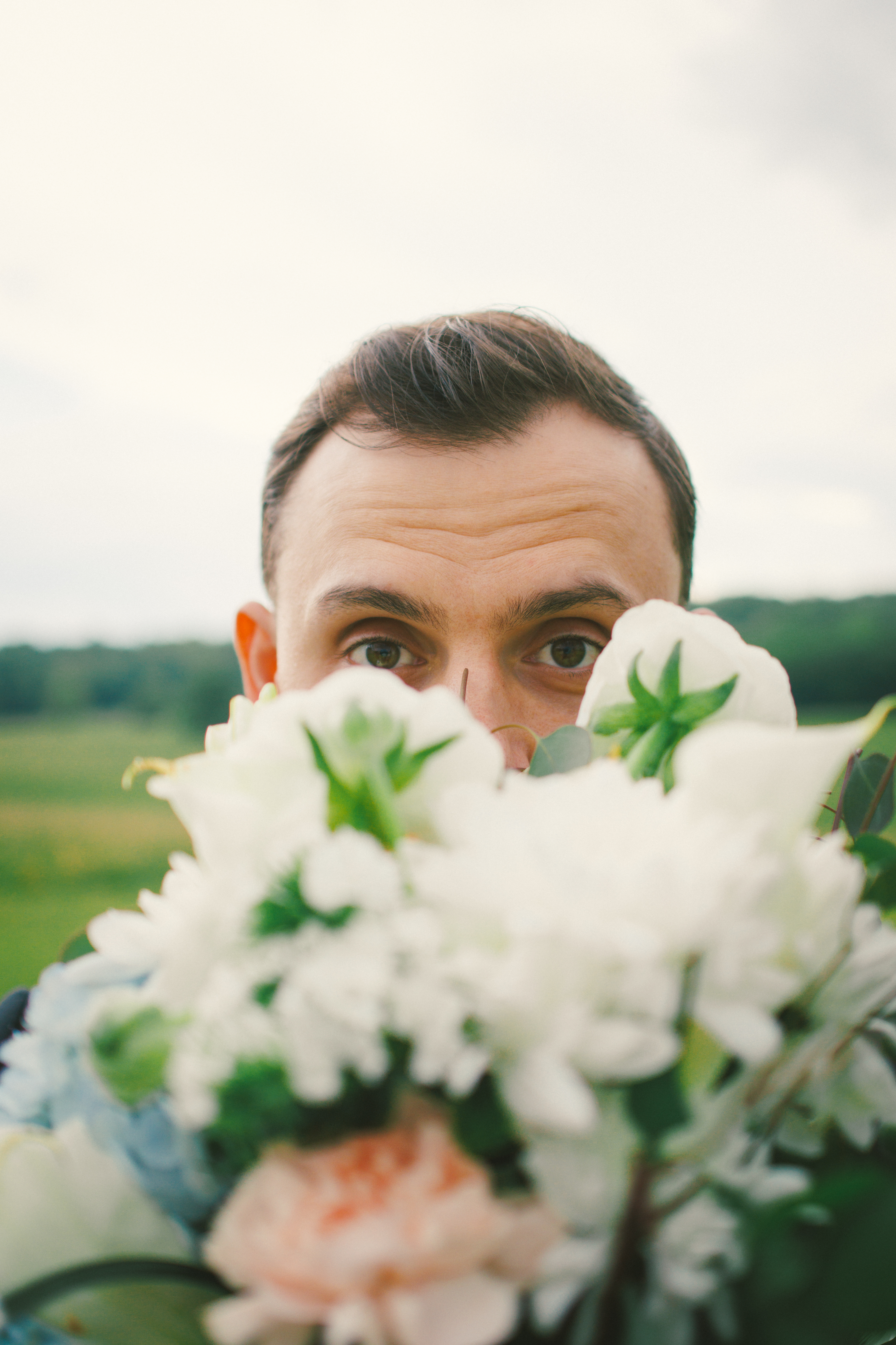 Groom peeking over a bouquet of white flowers outdoors on a cloudy day