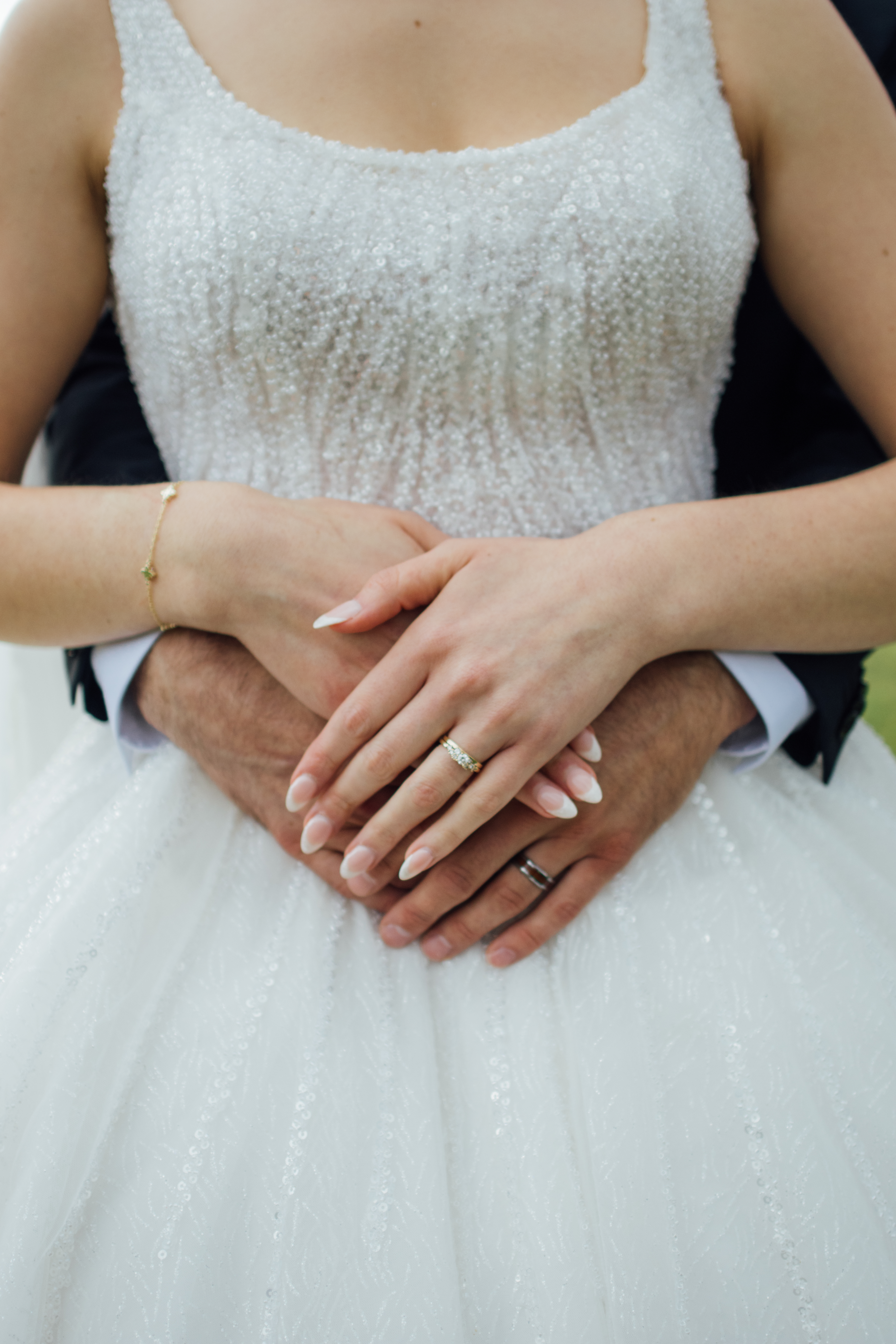 Close-up of a bride and groom's hands clasped, showing wedding rings and a beaded dress