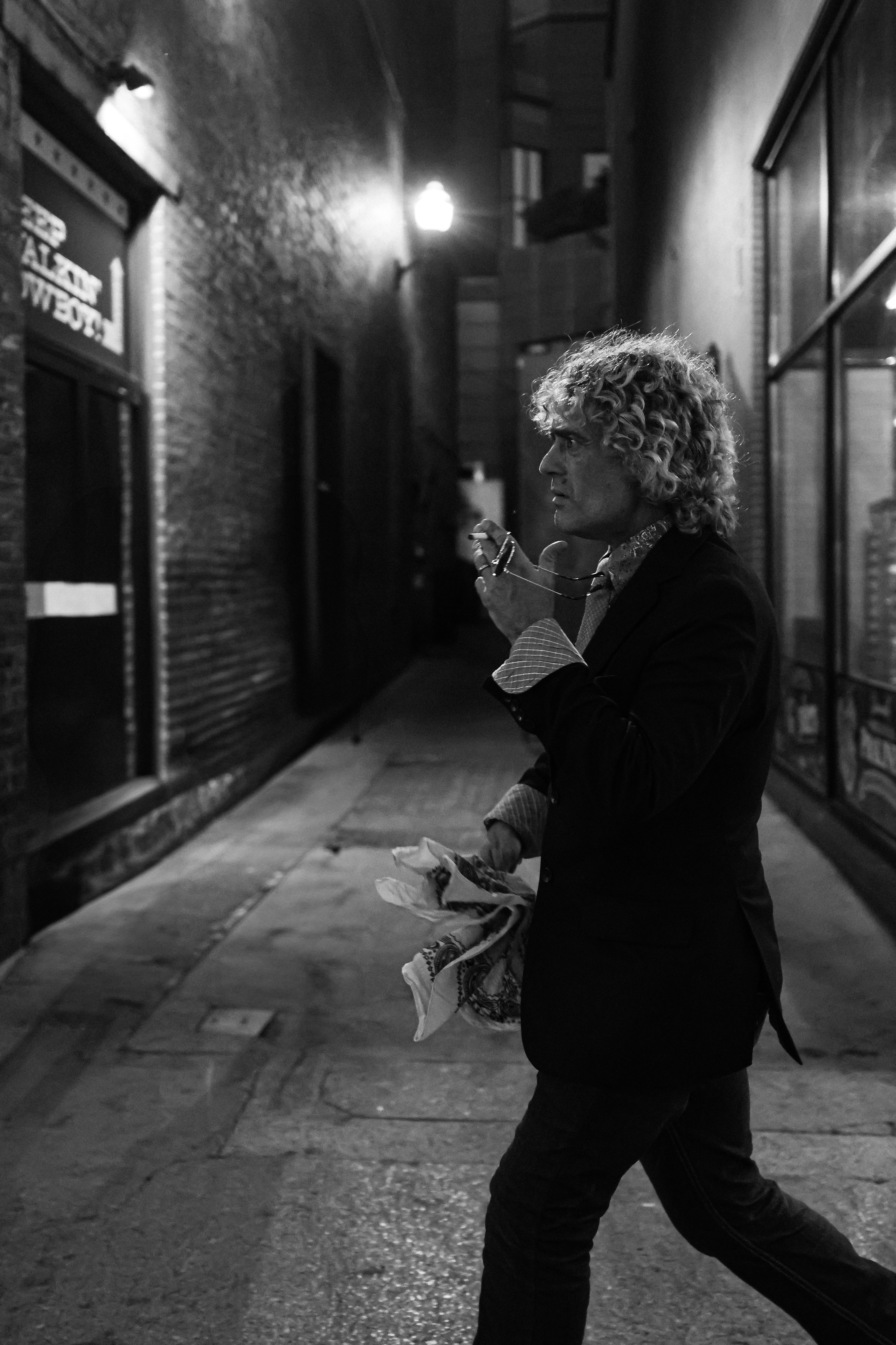 Man with curly hair walks down a dark alleyway holding a patterned scarf