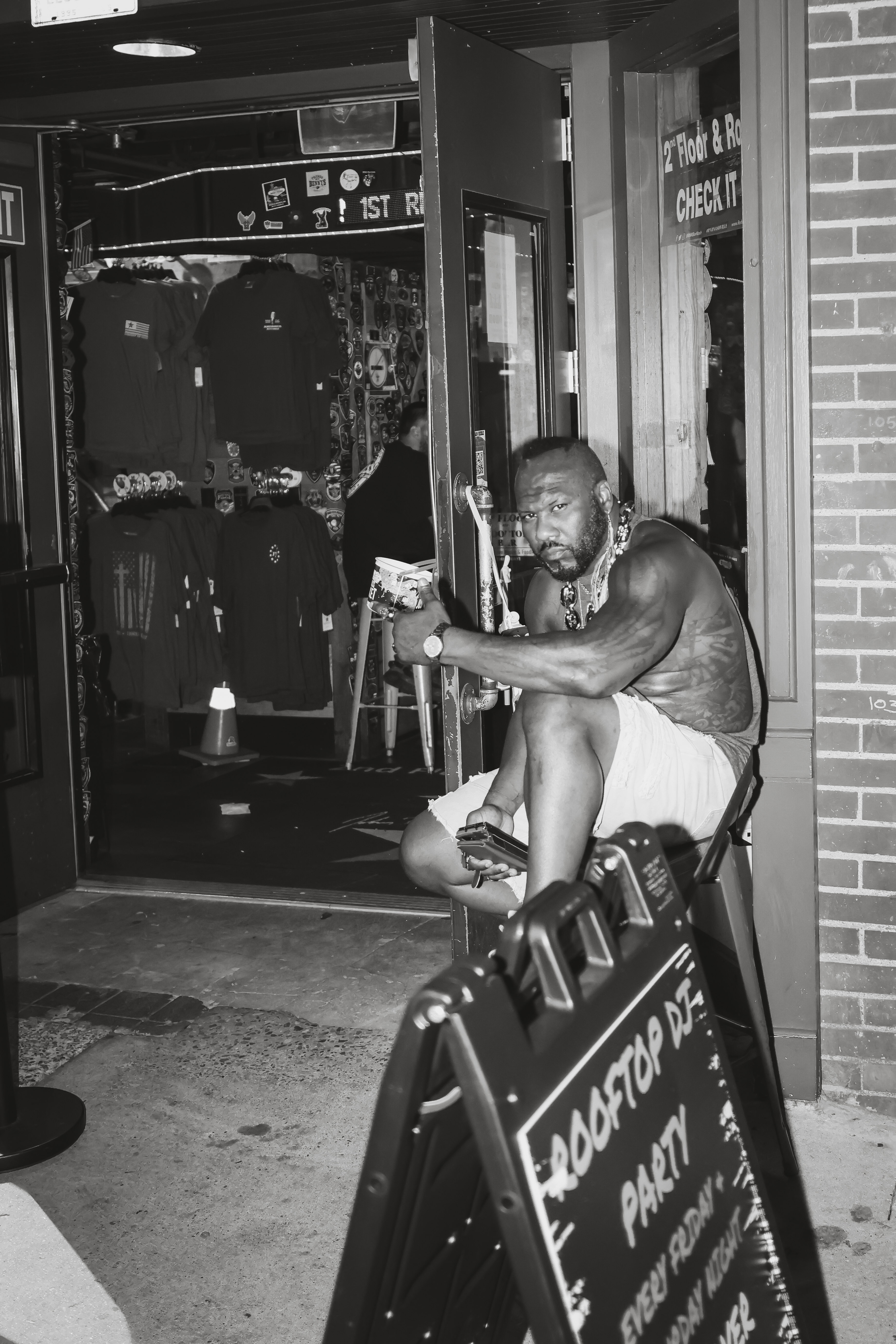 Man with tattoos and jewelry sits on a chair outside a store at night