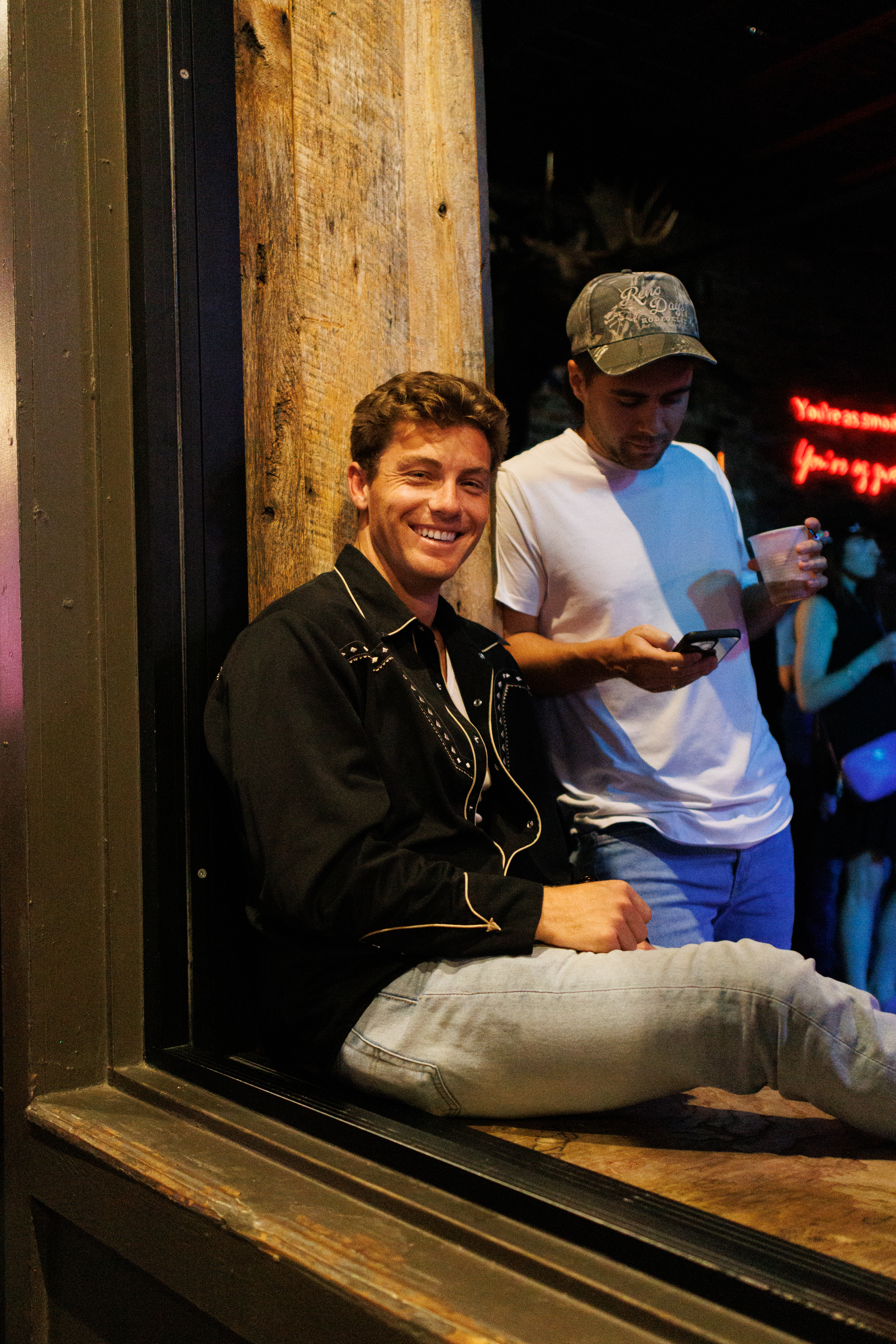 Man in black western shirt smiles sitting by window with friend holding drink