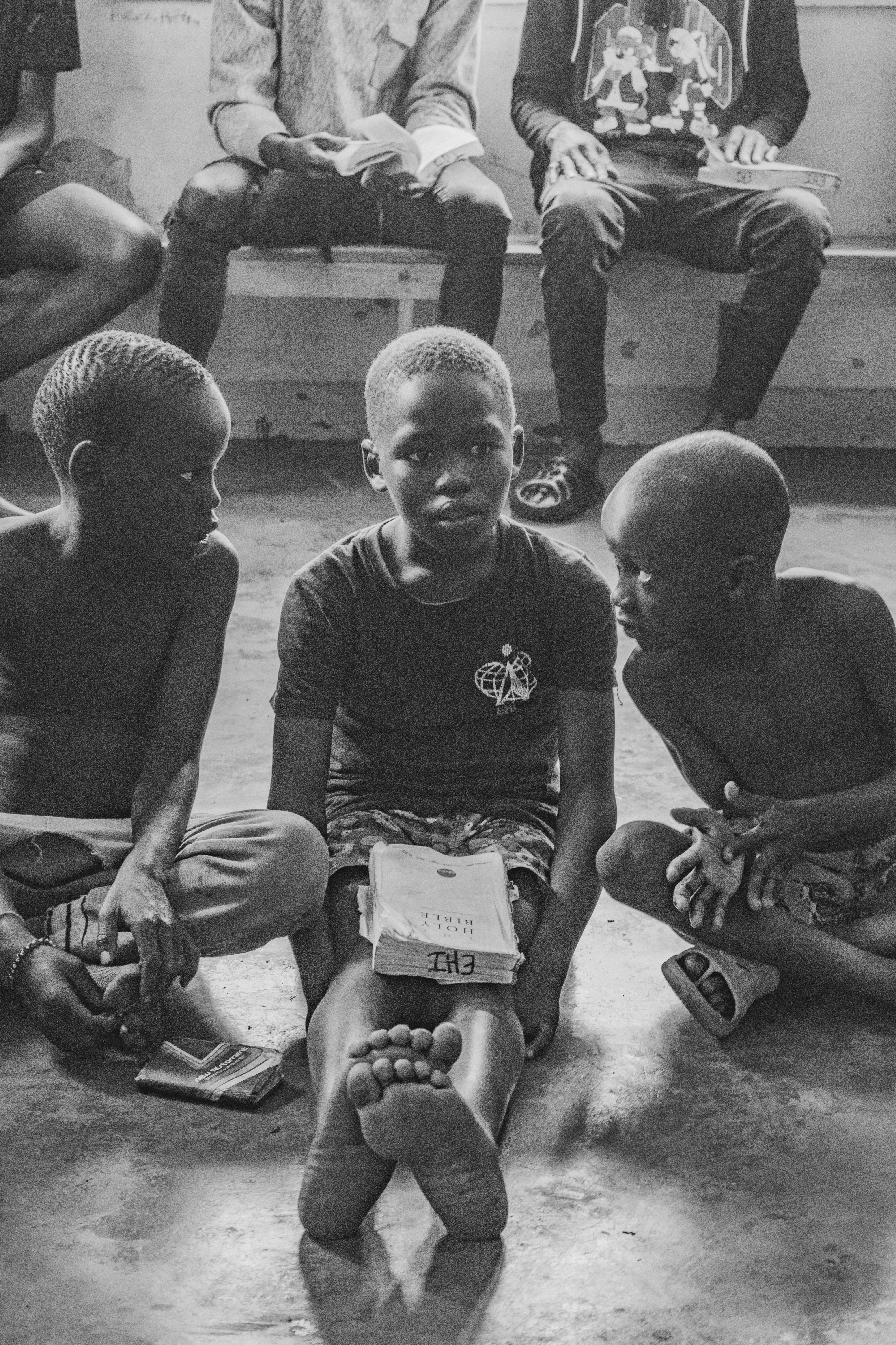 Three boys sit on the floor, one holding a Bible, in a candid street scene