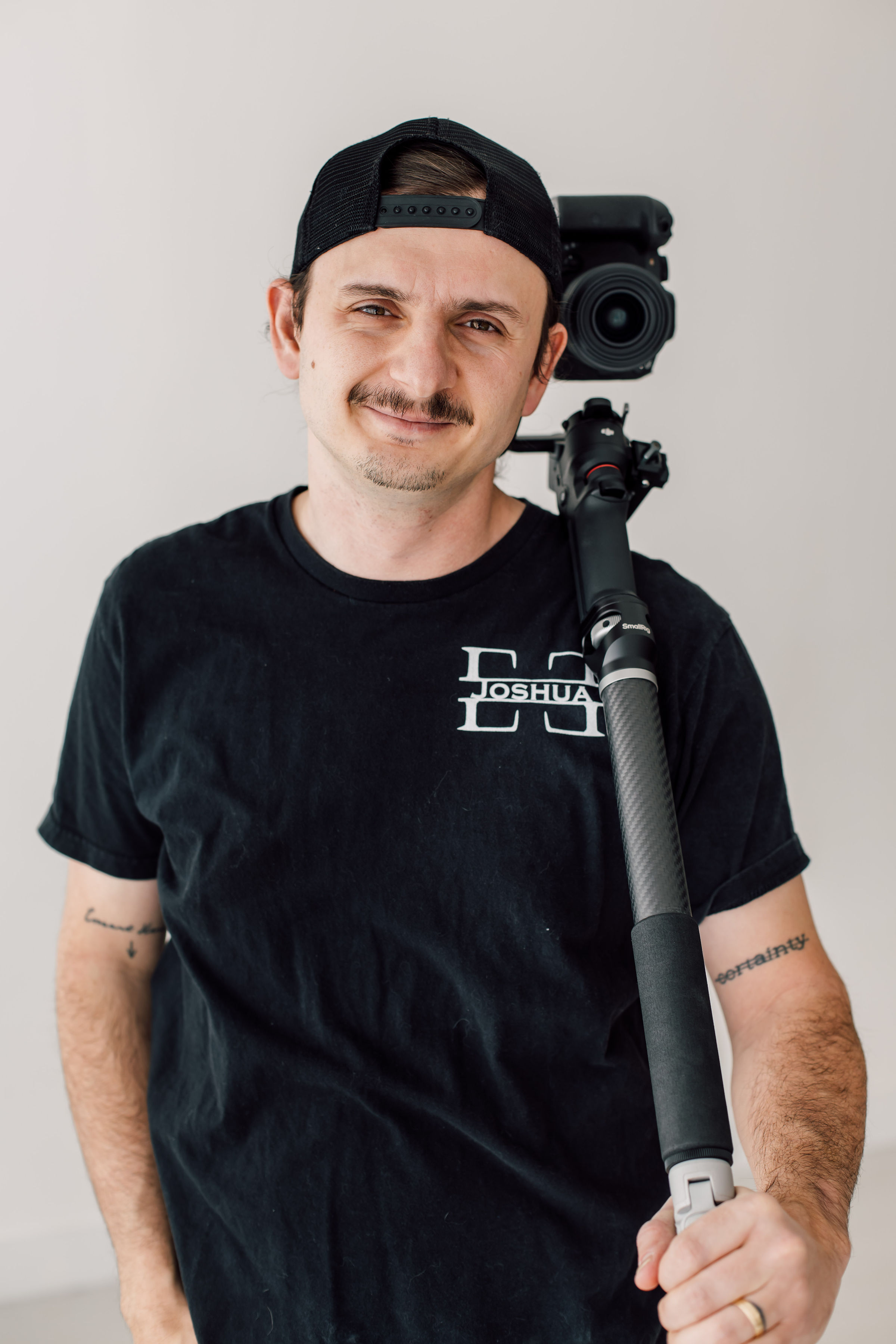 Videographer with camera rig smiles confidently against a light background