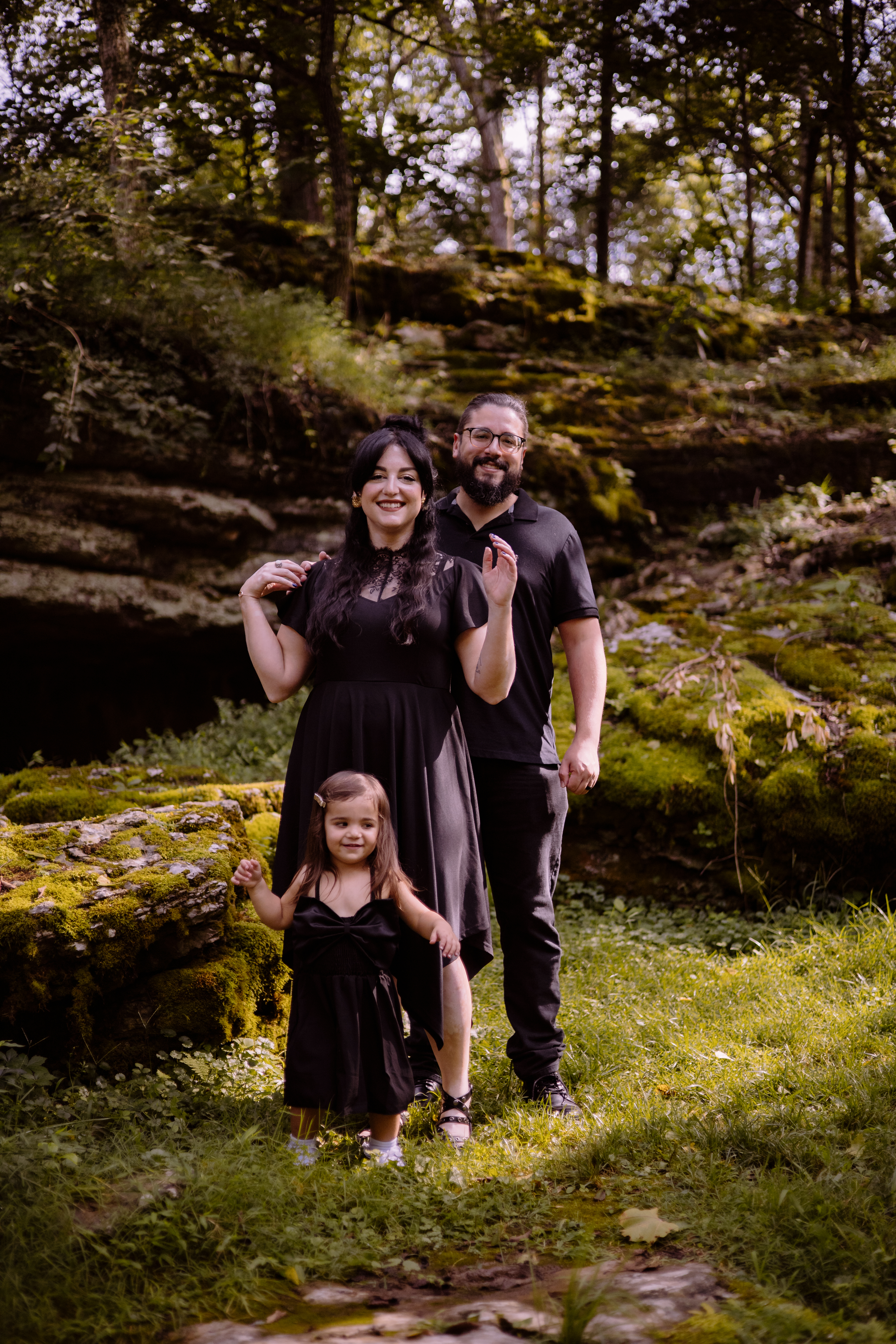 A smiling family of three poses in a mossy forest setting during a portrait session