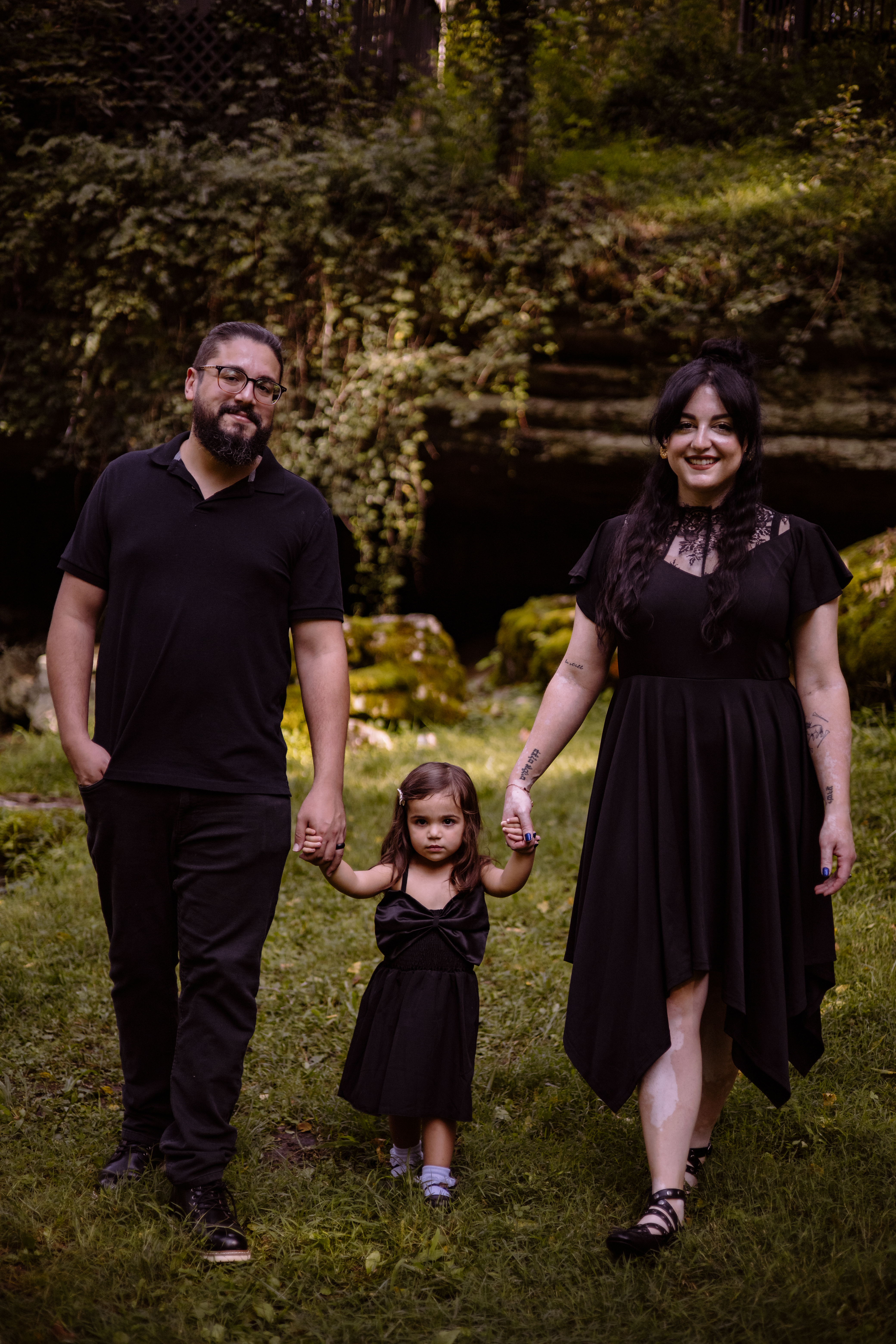 Family in black outfits walks hand-in-hand through a lush, green forest setting
